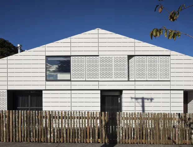 Front exterior view of a low-rise contemporary building with a prominent symmetrical gabled roofline and white horizontally articulated cladding. Decorative perforated screens flank the upper windows, while dark entry doors and windows contrast with the pale facade. A timber picket fence runs across the foreground under a clear blue sky.