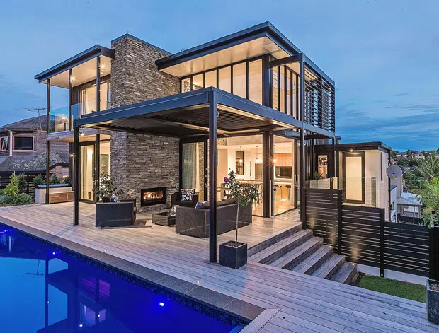Contemporary multi-level residence viewed from the pool deck at dusk, featuring extensive floor-to-ceiling glazing, dark steel framing, stacked-stone cladding, broad timber decking, and covered outdoor living areas. A linear outdoor fireplace, lounge seating, dining area, internal kitchen, and illuminated swimming pool are visible, with neighbouring homes and a landscaped hillside beyond.