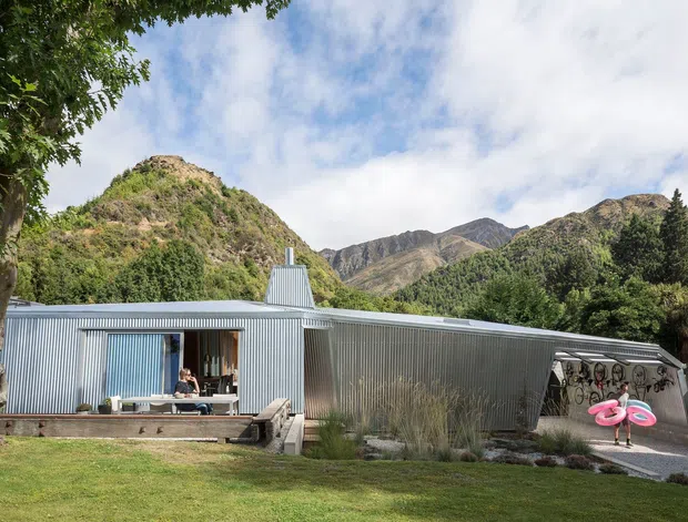 Exterior view of a low-profile contemporary home clad in corrugated metal, set against dramatic mountain terrain. The building has a long linear form, extensive glazing, a raised timber deck, and a covered outdoor area with playful bicycle artwork. Mature trees and lawn frame the foreground.