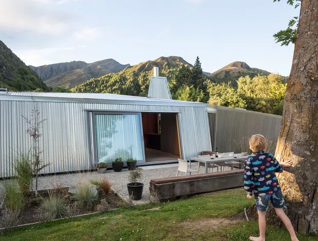 Single-storey corrugated metal-clad dwelling set against a mountainous landscape, with a low-pitched roof, large glazed sliding door, and a simple outdoor terrace containing a dining table and bench. Gravel landscaping, ornamental grasses, a lawn, and a mature tree frame the foreground.