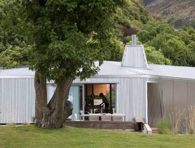 Single-storey contemporary house set in a lush, mountainous landscape. The building has extensive light-grey corrugated metal cladding and roofing, a low horizontal profile, and a central raised roof feature with a chimney or vent. Large glazed sliding doors open onto a timber deck and covered outdoor seating area. Mature trees and ornamental grasses frame the facade.