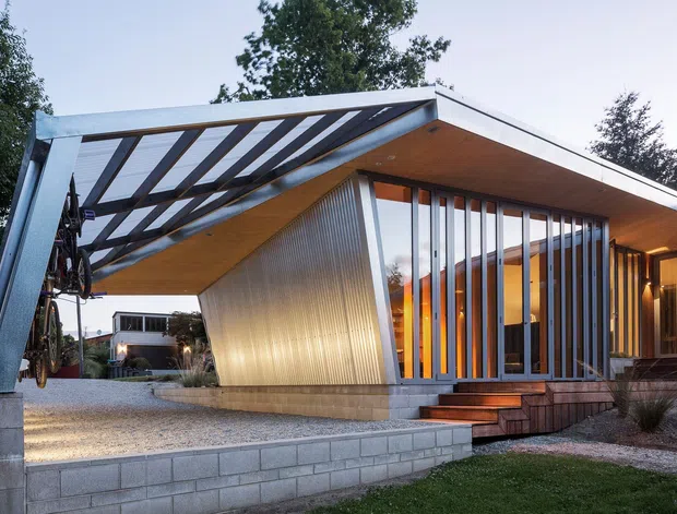 A striking modern single-storey residence viewed from the front, featuring a dramatic low-pitched projecting roof and open carport-like canopy supported by exposed steel framing. Full-height glazing is screened by closely spaced vertical timber or metal fins, while warm timber soffits and illuminated interiors create a welcoming contrast with the grey structural elements. The home sits on a slightly raised landscaped site with concrete block retaining walls, gravel paving, timber steps, and mature trees around it.