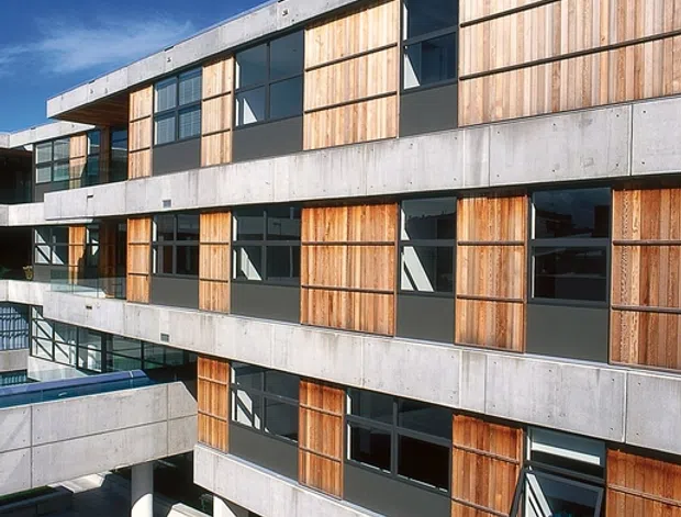 Exterior view of a multi-storey modern building with a strong grid of exposed concrete bands, vertical timber facade panels, and dark-framed windows. Recessed balconies and covered ground-level circulation frame a paved courtyard, with a blue sky visible above.