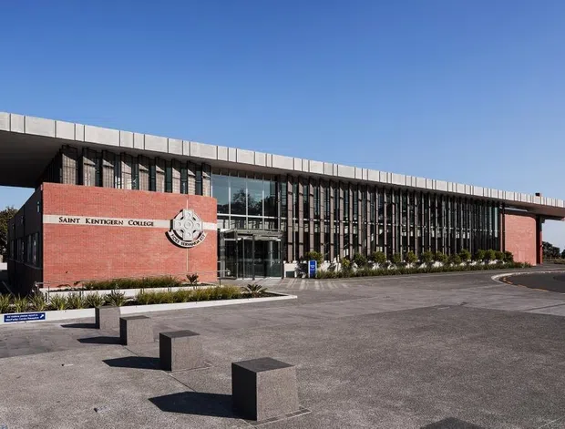 Exterior view of a contemporary educational building identified by signage as Saint Kentigern College. The long low-rise structure combines red brick end walls with an expansive glazed central facade screened by regularly spaced vertical fins, beneath a broad projecting flat roof. A paved forecourt with robust rectangular seating blocks occupies the foreground.