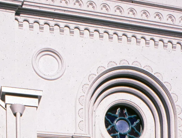 Close-up exterior view of an ornate pale rendered masonry facade, featuring a large arched window with decorative concentric mouldings, circular relief details, patterned friezes, and a steep gabled section. The architecture has strong historic ecclesiastical character, photographed against a deep blue sky.