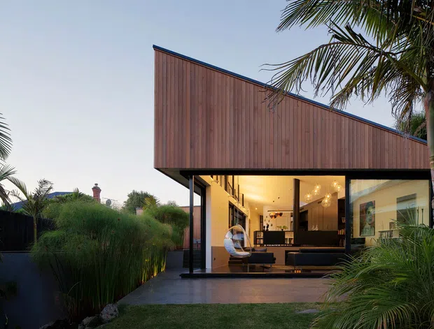 Contemporary single-storey pavilion-style home with a dramatic sloping timber-clad roof volume, expansive glazing, and an open living area visible through the facade. The building is set within lush tropical landscaping, with palms and ornamental grasses framing a concrete terrace and garden approach. Warm interior lighting contrasts with the dark exterior setting.