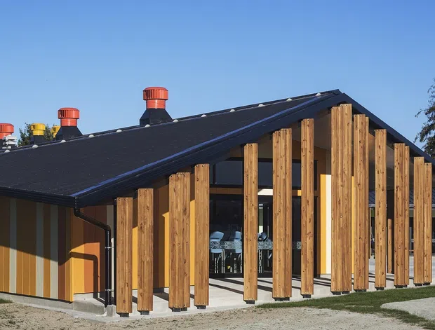 Exterior view of a contemporary single-storey building with a dark pitched gable roof and a striking timber-screened facade. Vertical timber posts create a covered veranda-like edge around extensive glazed openings, while the side wall combines warm orange and muted grey vertical cladding. Several rooftop vents are visible, with a paved entry path and lawn in the foreground.