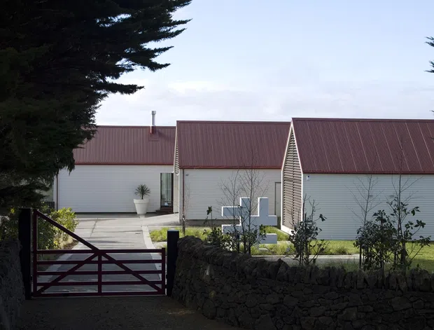 Exterior view of a low-rise contemporary building complex with long corrugated metal roofs, pale horizontal-clad walls, and a dark metal driveway gate. A rough stacked-stone retaining wall and landscaped garden sit in the foreground, with mature evergreen trees framing the property.
