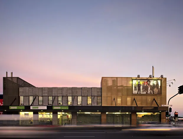 Twilight exterior photograph of a contemporary multi-storey commercial building on a prominent urban street corner. The facade combines dark perforated metal screens, warm brown brick or masonry panels, extensive ground-floor glazing, rooftop mechanical elements, and a large illuminated advertising or display panel. Traffic signals, street lighting, and blurred roadway traffic reinforce the urban commercial context.