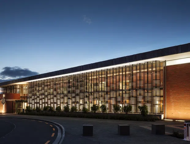 A contemporary institutional building photographed at dusk, with a long low roofline, extensive floor-to-ceiling glazing, warm illuminated interiors, dark vertical screening, and broad red-brick wall sections. The paved forecourt includes low bollards and a curved roadway.