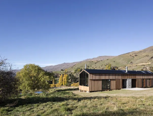 A low, single-storey contemporary house set in a dry, open mountain valley landscape. The building has dark timber-clad walls, a simple low-pitched roof, large glazed end wall, and a covered or recessed entry area. Sparse trees and rolling, rugged hills surround the dwelling under a clear blue sky.