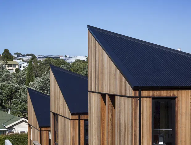 Exterior architectural view of a row of narrow, stepped timber-clad building volumes with steeply pitched dark metal roofs. The repeated gabled forms descend along a sloping site, with mature vegetation and suburban buildings in the background under a clear blue sky.