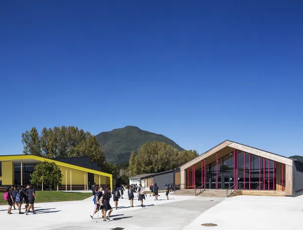 Exterior architectural view of a contemporary educational or community campus, with low-slung buildings arranged around a broad paved forecourt. The main gabled building has extensive glazing and distinctive bright pink vertical framing, while the adjacent building has vivid yellow cladding and dark glazing. People are present for scale, with a wooded mountain rising behind the site under a clear blue sky.
