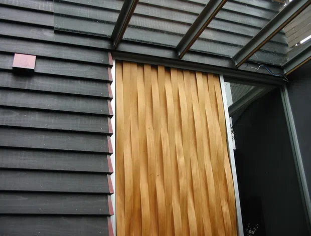 Close-up exterior architectural detail showing dark horizontal weatherboard cladding beside a tall vertical timber feature door or wall panel. A translucent corrugated roof canopy with dark structural framing covers the area, and a dark-framed glazed opening is visible at right. The image emphasizes contrasting cladding materials and entry detailing.