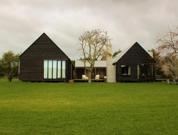 A low, modern residential composition set in a broad lawn, with two dark timber-clad pavilion-like buildings featuring steep triangular gable roofs. Large glazed openings connect the buildings to a central pale masonry volume and covered outdoor area. Mature deciduous trees and an overcast sky surround the house.