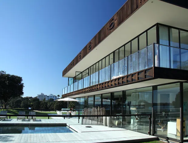 Contemporary multi-level residence with extensive floor-to-ceiling glazing, projecting upper-level balconies, dark timber soffit and fascia detailing, and a broad pale terrace surrounding a rectangular swimming pool. The house overlooks landscaped grounds and appears bright, open, and strongly connected to the outdoors.