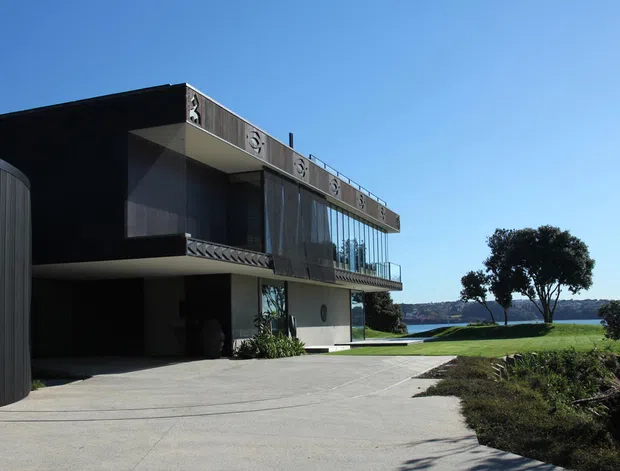 Modern multi-level waterfront residence viewed from the driveway, with a bold cantilevered dark upper volume, extensive glazing, projecting balcony, curved vertical-panel garage or entry enclosure, and a broad concrete forecourt. The landscaped site slopes toward visible water beyond the house.