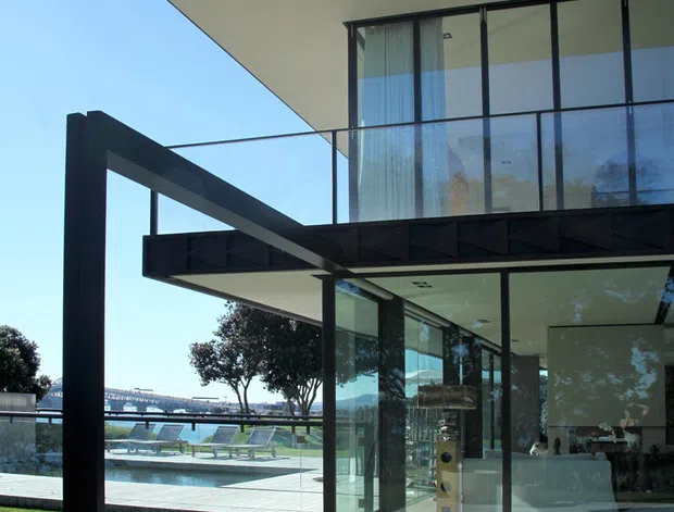 Exterior view of a contemporary multi-level residence with strong horizontal cantilevered forms, dark structural framing and fascia, extensive floor-to-ceiling glazing, and a glass balustrade. A tiled terrace with potted plants overlooks a landscaped lawn and swimming pool, with a distant waterfront setting visible beyond. The interior living area is visible through the glazing.
