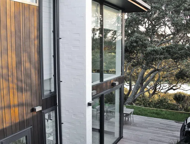 Exterior view of a contemporary multi-level home with dark vertical timber cladding, white-painted brickwork, black-framed floor-to-ceiling glazing, and a timber deck overlooking mature trees. A barbecue is positioned on the deck.