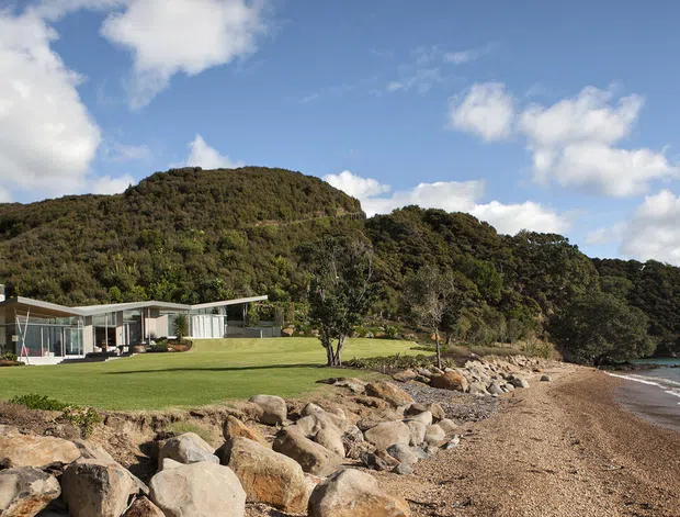 A low-profile contemporary house set directly beside a rocky beach and calm blue water, backed by densely vegetated hills. The home has extensive floor-to-ceiling glazing, pale horizontal roof planes, and a broad manicured lawn extending toward the shoreline. The scene is photographed in bright daylight with scattered clouds and a natural coastal setting.
