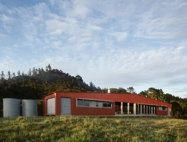A low, elongated single-storey building set in a grassy rural landscape, with a strongly horizontal form, red corrugated metal cladding, broad dark-framed glazing, and roller doors at one end. Large cylindrical tanks sit beside the building, while wooded hills rise behind it under a partly cloudy sky. The image is an exterior architectural photograph with a restrained contemporary utilitarian character.