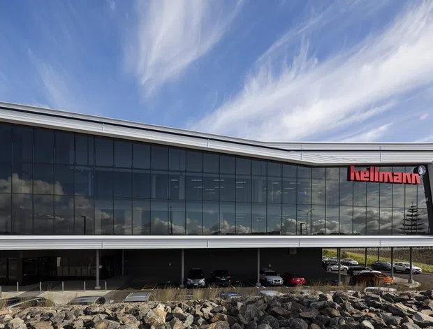 Exterior view of a contemporary commercial building with a long, gently curved roofline, extensive dark reflective curtain-wall glazing, and a sheltered ground-level parking area. The red Hellmann logo is displayed on the upper facade. The building sits beside a landscaped rocky embankment under a bright sky.