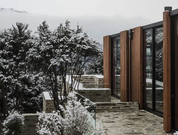 A modern mountain-side exterior or terrace in a snowy landscape, featuring a low stepped stone retaining wall and paving leading alongside a dark-framed glazed facade with vertical timber-clad sections. Snow-covered trees and a distant mountain are visible beyond the building. The design has a restrained contemporary character with strong natural stone, timber, and black metal glazing.