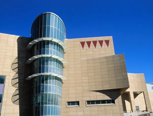Exterior view of a contemporary multi-storey commercial building with a prominent cylindrical glazed stair or circulation tower, curved metal sunshade platforms, and large beige rectangular wall planes. The facade includes narrow horizontal windows, geometric projecting volumes, and red triangular decorative panels beneath a clear blue sky.
