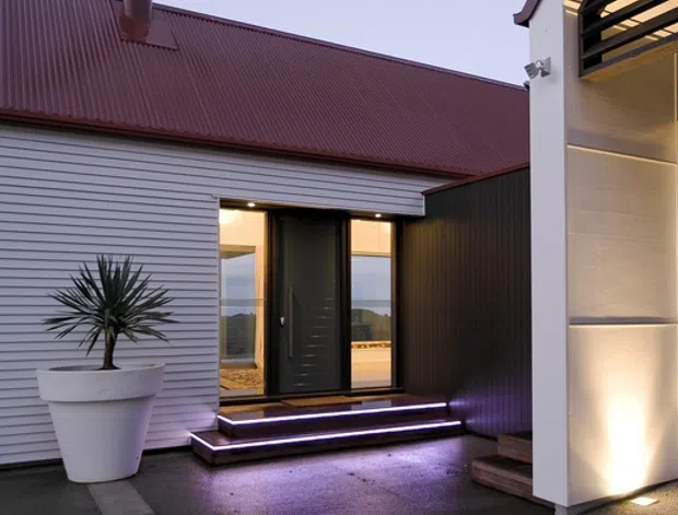 Twilight exterior view of a contemporary residential entry courtyard. The facade combines horizontal silver-grey metal cladding with a dark corrugated metal wall and a steep red-burgundy corrugated roof. A glazed entry door is reached by illuminated steps, while the adjacent white rendered structure has warm recessed lighting and a dark horizontal louvre screen. Dark paving, a large white planter, and a sculptural spiky plant complete the scene; distant landscape is visible through the glazing.