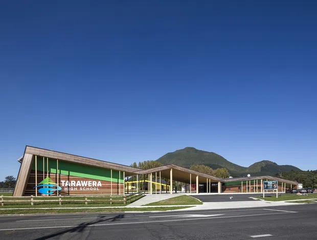 Exterior view of a low-rise school campus with long, angular timber-clad wings, extensive glazing, covered walkways, landscaped lawns, and a prominent Tarawera High School sign. A mountainous landscape forms the background beneath a clear blue sky.