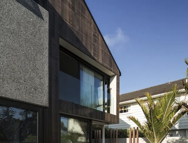 Exterior view of a contemporary two-storey residential extension or townhouse, featuring a steep gabled form clad in dark vertical timber beside a textured grey concrete wall. Large glazed openings connect to a paved courtyard terrace with outdoor dining furniture, screening battens, and a palm tree. A neighbouring weatherboard-style house is visible in the background under a clear blue sky.