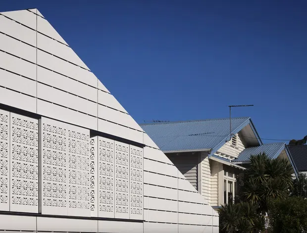Exterior architectural photograph showing a sharply angled white-clad building facade with horizontal panel lines and a large patterned breeze-block screen. Behind it is a neighbouring light weatherboard house with pitched corrugated metal roofs, set against a clear deep-blue sky and subtropical planting.