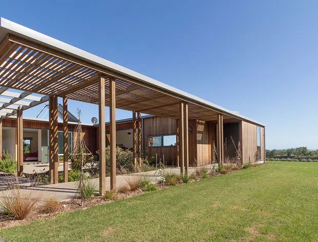 A low, single-storey contemporary building viewed from the landscaped exterior, featuring warm timber-clad walls, a broad flat roof, and a large timber pergola with slatted screening over a covered outdoor walkway or courtyard. The building opens onto a lawn and planted garden, with strong horizontal lines and abundant sunlight.