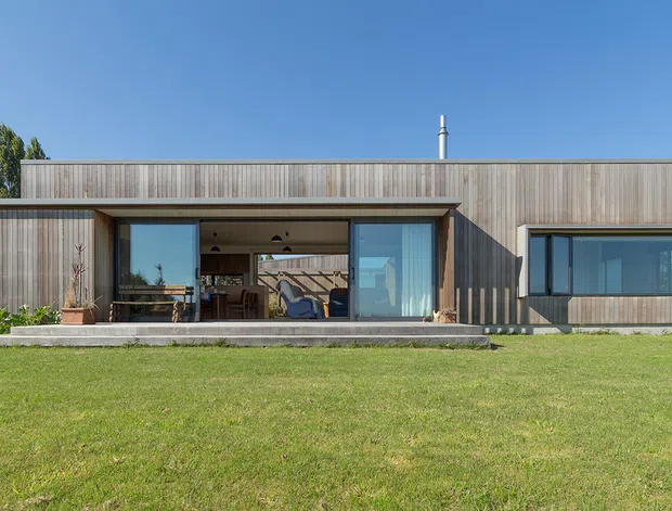 Front exterior view of a low, single-storey contemporary house set behind a broad lawn. The building has a simple rectilinear form with extensive floor-to-ceiling glazing, a recessed covered outdoor living area, and vertical timber cladding in natural grey-brown tones. A flat roof with a projecting edge and minimal detailing reinforces the modern design.