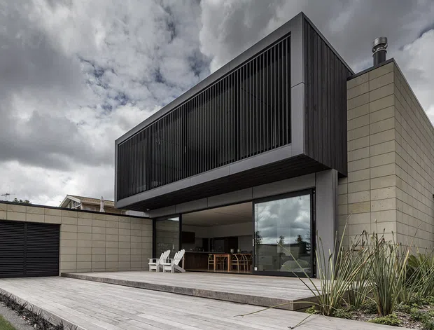 Contemporary single-storey residence with a strong geometric composition, combining pale masonry walls with a projecting dark upper volume screened by vertical slats. Large glazed openings connect the interior to a broad timber deck, while a separate garage sits to the left. The image is an exterior architectural view under an overcast sky, with restrained coastal-style landscaping in the foreground.