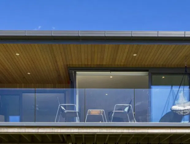 Exterior view of a contemporary elevated building with a deep recessed balcony or terrace. The facade combines extensive floor-to-ceiling glazing, transparent glass balustrades, dark metal framing, and a warm timber-lined soffit and ceiling. Minimal recessed downlights and outdoor seating are visible, with blue sky reflected in the glass.