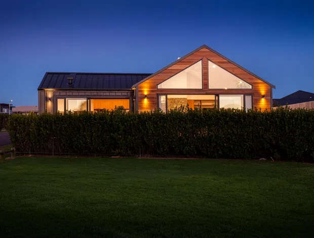 A single-storey suburban house photographed at dusk, viewed from across a neatly clipped hedge and lawn. The home has a prominent front-facing gable with warm timber cladding, large glazed openings, a dark metal roof, and a lower side wing with additional glazing. Interior lighting creates a warm glow against the deep blue evening sky.