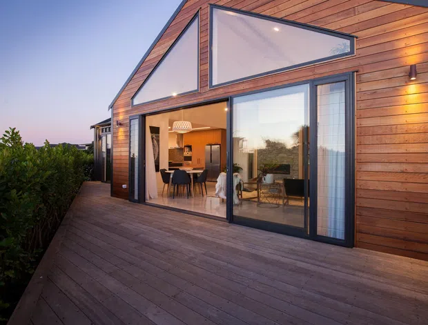 Exterior view of a contemporary timber-clad home at dusk, with a broad timber deck, large black-framed glazed doors and windows, and prominent angular clerestory glazing beneath a pitched gable roof. A warm, illuminated dining and kitchen area is visible through the glazing.
