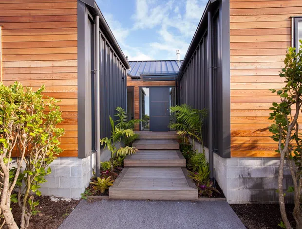 Exterior view of a contemporary single-storey dwelling entrance, framed by two timber-clad wings with dark metal vertical cladding and trim. A timber boardwalk and steps lead through a planted entry courtyard to a dark front door beneath a low metal roof. Concrete block foundations, glazing, and landscaped planting are visible.