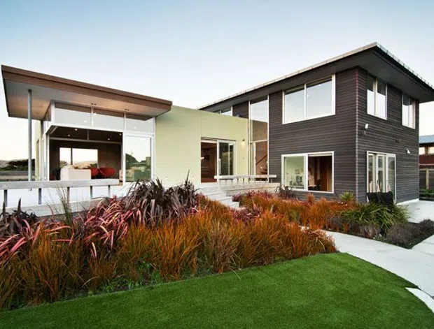 Contemporary two-storey residential exterior with a low-pitched roof, dark horizontal cladding, pale rendered walls, extensive glazing, and a covered outdoor living area. The house is set within a landscaped front garden featuring ornamental grasses and a paved walkway.