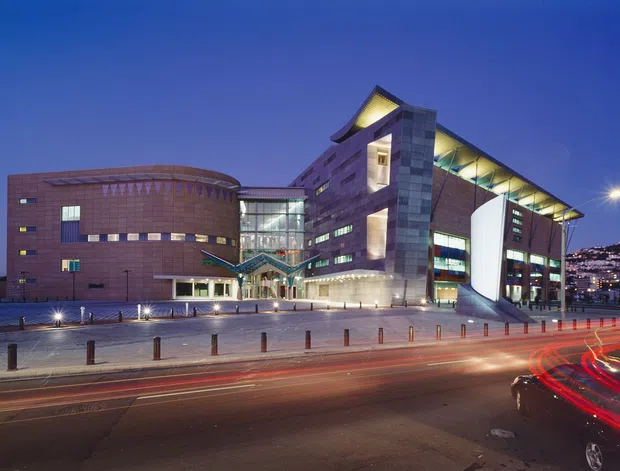 Night-time exterior view of a large contemporary civic or commercial building beside a busy road. The complex combines curved red-brick volumes with a taller angular glazed and metal-clad central section, illuminated roof overhangs, extensive windows, and an open paved forecourt. Traffic light trails and a hilly, illuminated urban backdrop reinforce the city setting.