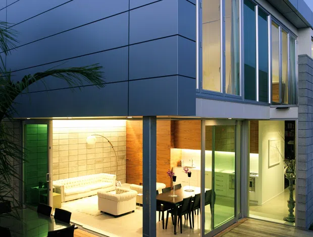 Twilight exterior view of a sleek multi-level contemporary home with dark grey panelled cladding, extensive floor-to-ceiling glazing, and a timber deck. Warmly illuminated living and dining areas are visible through the glass, with a kitchen further inside. The composition highlights the building facade, corner glazing, and indoor-outdoor connection.