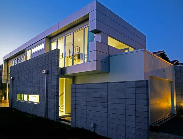 Contemporary multi-level residential facade photographed at dusk, featuring dark grid-pattern masonry walls, smooth grey panel cladding, projecting cubic volumes, large illuminated windows, and a recessed entry. Warm interior lighting contrasts with the cool blue exterior light.