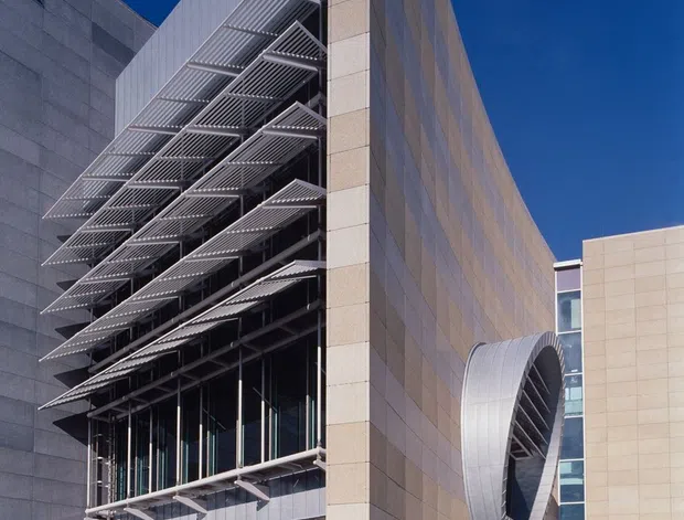 A dramatic close-up exterior view of a large contemporary institutional or commercial building. Tall angular beige stone-clad walls frame a recessed glazed facade with layered horizontal metal sunshade fins, while a curved silver architectural canopy projects from the side. Reflective dark water and broad steps occupy the foreground under a deep blue sky.