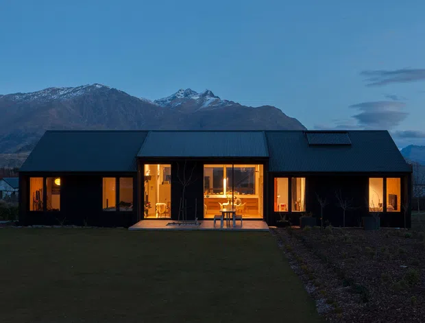 A single-storey contemporary house photographed at dusk, set against a dramatic snow-dusted mountain range. The dark metal-clad facade has a long pitched roof, warm amber light glowing through large windows and glazed doors, and a central recessed entry or living area opening toward a lawn and cultivated garden.