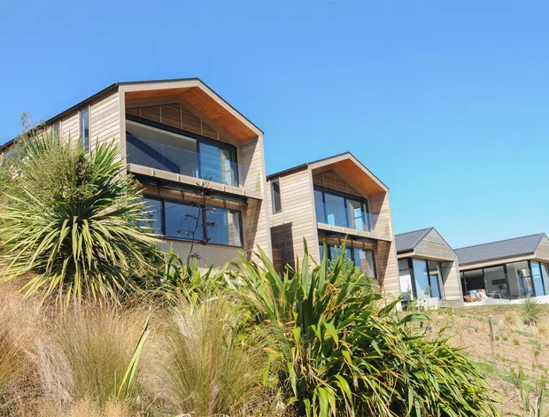 Exterior view of a row of contemporary multi-level homes on a sloped, landscaped site. The dwellings feature timber-look horizontal cladding, prominent glazed openings, projecting upper-level balconies or recessed terraces, and steeply pitched gable-like roof forms. Native grasses and spiky plants occupy the foreground.