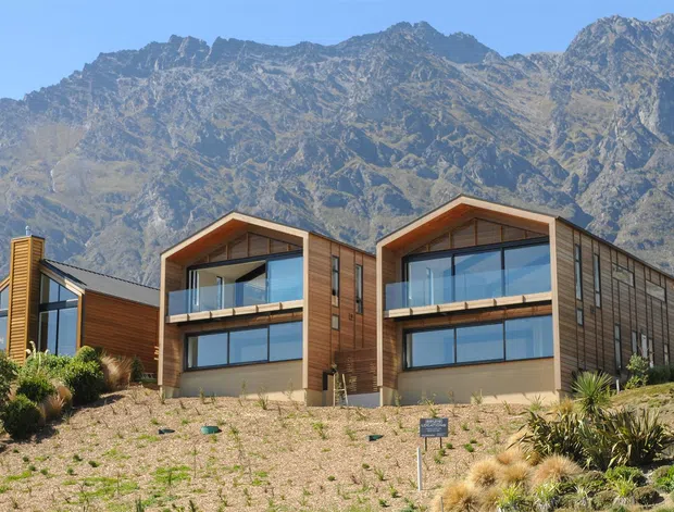Exterior view of a contemporary multi-unit residential development set against dramatic rugged mountain terrain. The buildings feature steeply angled gabled roof forms, extensive glazing with clear balustrades, warm timber cladding, dark metal roofing, and landscaped rocky foregrounds.