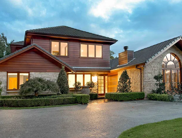 Exterior view of a large multi-wing residence at dusk, featuring dark tiled pitched roofs, horizontal timber weatherboard cladding, pale stone walls, warm illuminated windows and doors, and carefully clipped hedges and topiary around a broad paved driveway. A prominent arched window and masonry facade add traditional character.