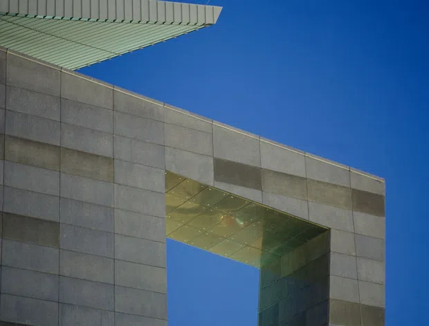Upward-looking architectural photograph of a modern institutional or commercial facade clad in large rectangular grey panels. A prominent rectangular void cuts through the facade, with a reflective metallic soffit and deep reveals, while a projecting ribbed roof or canopy extends at the upper left. The composition emphasizes geometric form, sharp edges, material joints, and contrast against a clear deep-blue sky.