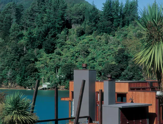 A modern multi-level house set beside a deep blue lake, backed by densely forested hills. The building combines warm timber cladding with dark charcoal rendered volumes, projecting roof forms, and strong vertical structural elements. Native-looking spiky planting and dense vegetation frame the foreground and right side of the image.
